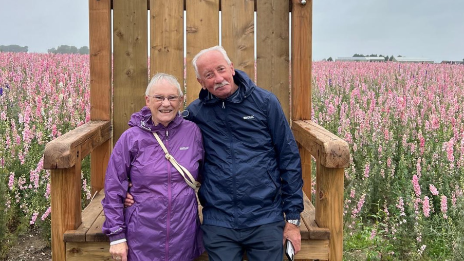 An older man and woman in rain jackets, stood in front of a field of flowers and a large wooden chair