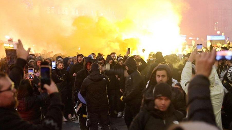 A crowd of people, many with dark jackets and hoods on, standing in front of a huge bright fire