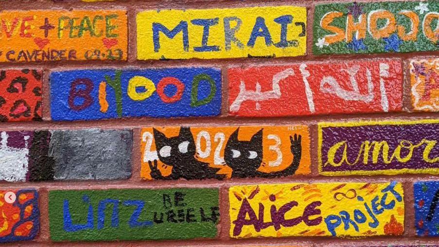 A close-up picture of the bricks on the wall. The bricks were originally red, but have been painted blue, red yellow, green, orange and purple. Members of the public have painted designs on them such as cats, their names, leopard spots, and the words 'love and peace'.