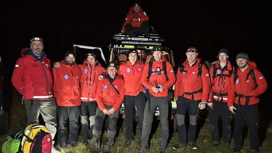 Nine of the rescuers stand in front of a land rover. Another team member is on top of the car. They all wear red jackets and black trousers. There are some backpacks in front of them on the left. It is dark. There are nine men and one woman in the team. 