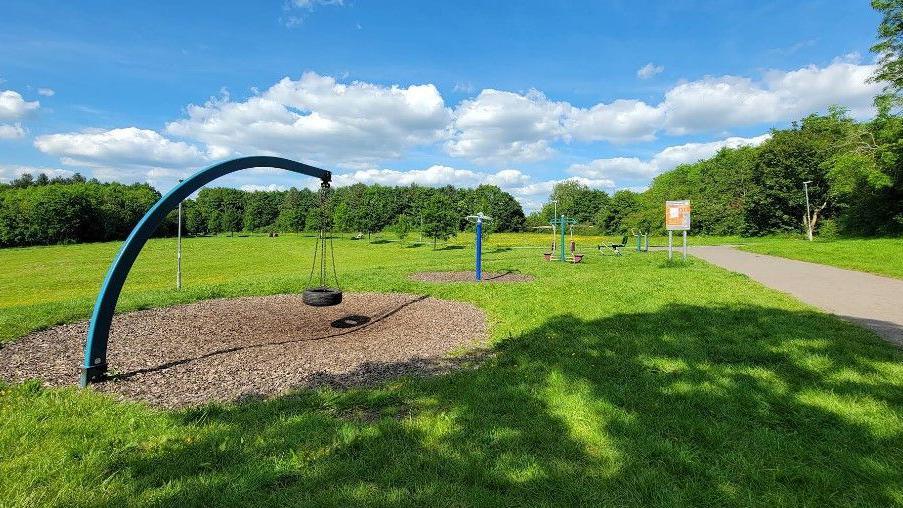An empty play park on a green with minimal play equipment. The image was taken on a sunny day and trees can been seen in the background.
