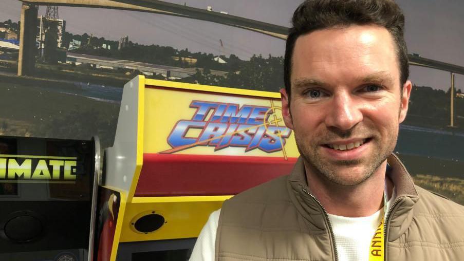 A man is smiling at the camera and he is standing in front of an arcade game. He is wearing a white top with brown Gillet.   