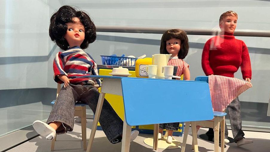 A Sindy doll sits at a 1960s-style table with a smaller female doll and a male doll