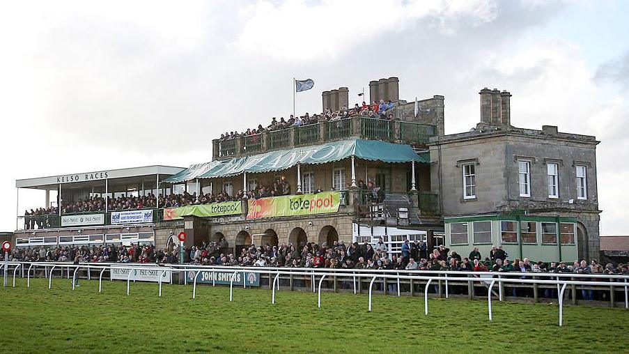 The main stand at Kelso racecourse with big crowds outside and a Scotland flag flying