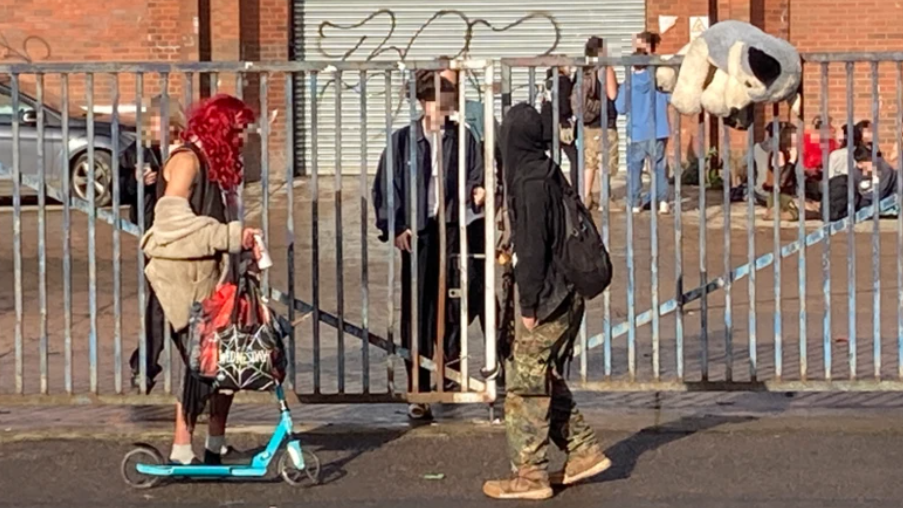 About a dozen youngsters milling about in daylight outside the former Wickes building with graffiti on a large garage entrance in the background and steel gates in the foreground