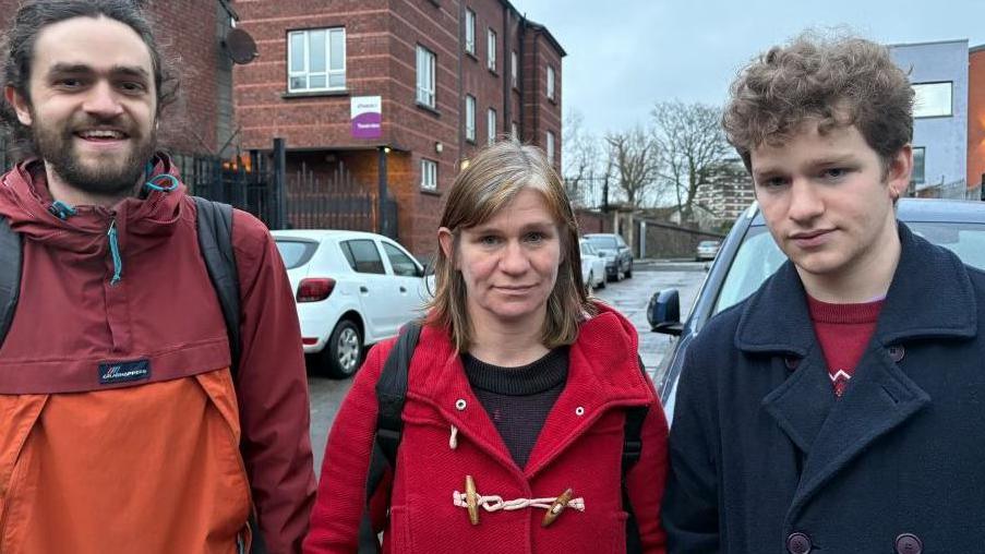 Two men and a woman in the middle. The man on the left has a beard and is wearing a red and orange top. The woman has blonde hair and is wearing a red coat with a black top underneath. The man on the right has curly brown hair and is wearing a dark coat with a red T-shirt underneath.