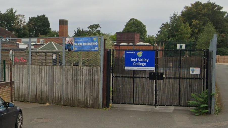 Large black gates and a wooden fence outside a school. There is a blue sign saying "Ivel Valley College" on the gate. There are brick buildings visible on the other side of the gate and trees behind.