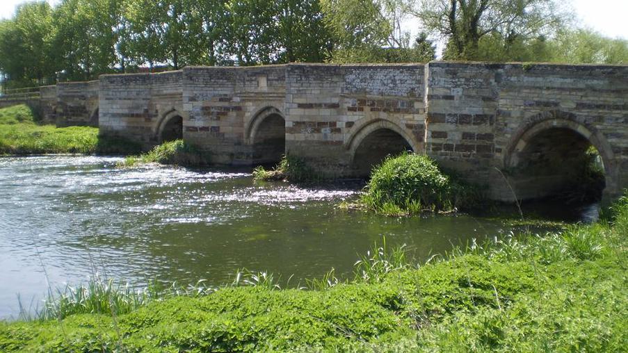 An ancient stone bridge with four arches over a river. It is a sunny day and water is flowing under the bridge. Some greenery can be seen on both banks of the river. There are trees behind the bridge.