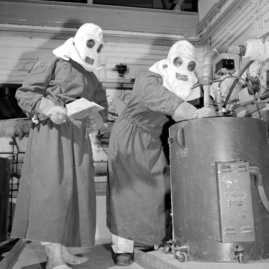 The two workers in the black and white image are wearing heavy overalls and masks with glass-covered eye holes. They are working at a metal drum.