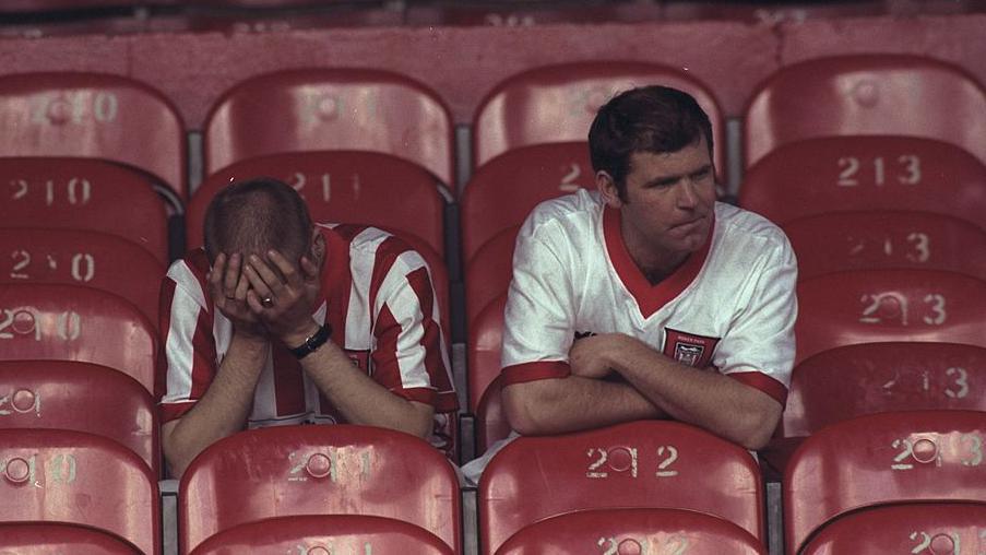 Two Sunderland fans remain seated at Selhurst Park. One, wearing a red-and-white home shirt, has his head in his hands. To his left, a fan wearing a white away jersey looks forlorn. They are surrounded by empty plastic seats in the stand.