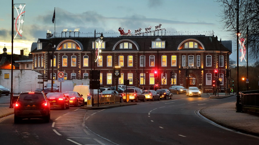 A grand, three storey building with arched windows on the top floor, pictured at twilight. There are vehicles queuing at traffic lights in front of the building, and a large model of reindeers and a sleigh can be seen on the building's roof.
