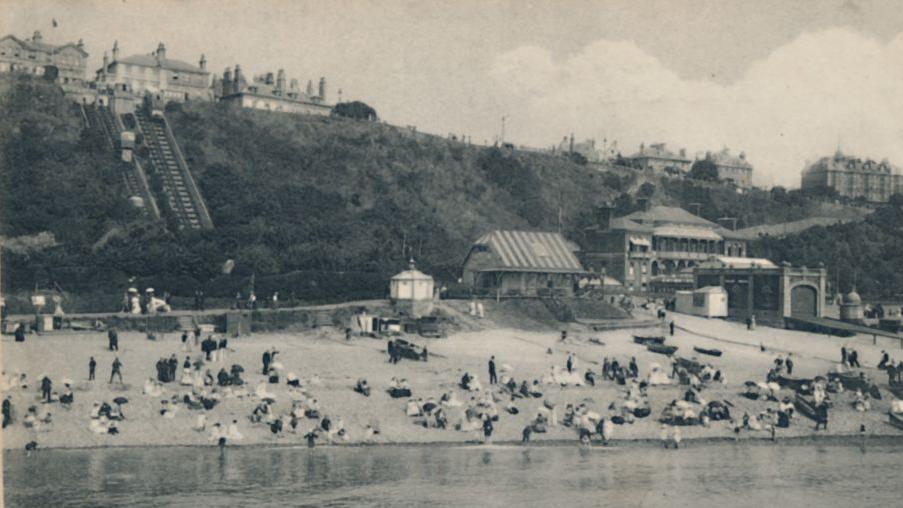 A black and white archive image of Edwardians at the beach. In the background is a funicular. 