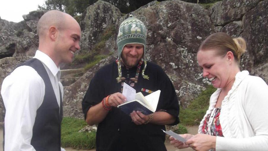 Matt and Gemma stand side on, with Gemma holding a piece of paper and Matt watching attentively, wearing a white shirt and grey waistcoat. Between them is a wedding officiant wearing a Peruvian hat.
