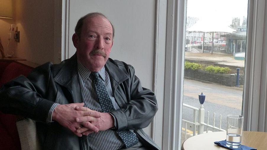 A picture of Alun Lloyd Jones looking into the camera. He is sat on a table on his own with a glass of water.