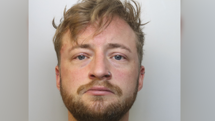 A police mug shot of a man with fair hairm blue eyes, a nose ring and a beard looking directly at the camera.