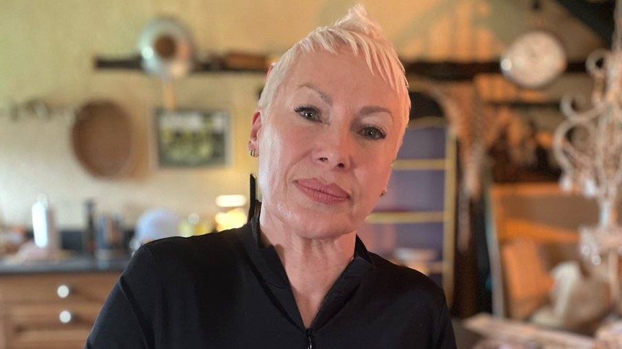 A head and shoulders image of a woman with very short peroxide blonde hair and wearing a black shirt, with a blurred background of a kitchen