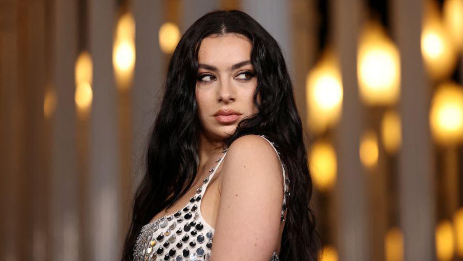 Charli XCX is standing sideways with her face turned towards the camera. She is looking off to the side and pouting. She is wearing a white top with large silver sequins and her long, black, wavy hair is falling down her back and over her far shoulder. 