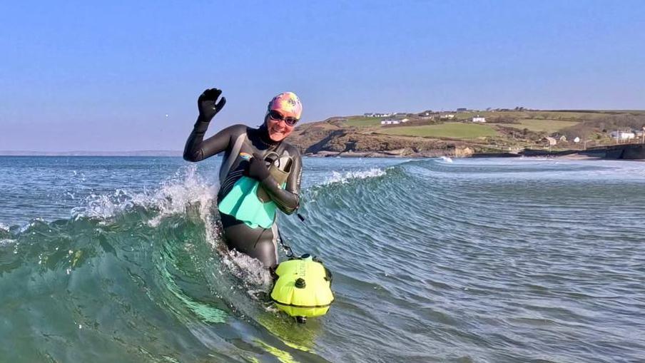Martina Morgan in the sea holding onto her floatation device and flippers. She is standing in a middle of a wave smiling. She is wearing a wetsuit, a swim cap and goggles.