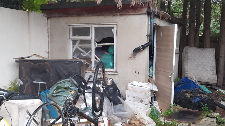 A back garden with a building in the back corner. It has a broken window and the door is ajar. In front of the building is an upside-down bicycle, broken furniture and debris. A dirty mattress is at the back of the garden leaning against a few tree trunks.