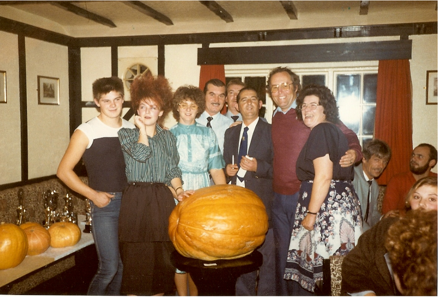 An old photo, shot and film, showing a group of men and women crowded in a dingey pub. They are surrounding a large pumpkin, placed on a wooden plinth.