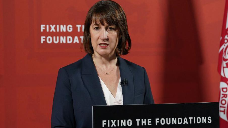 UK Chancellor Rachel Reeves standing in front of a red wall which reads fixing the foundations. She is wearing a navy blazer and white blouse