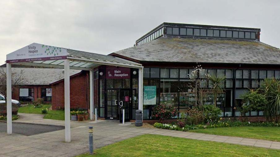 Street view image of the entrance to Trinity Hospice. It has a glass canopy leading over a flagged path leading to the main reception with grassed areas to the front. There are plants and flowers to the side of the building which has many windows