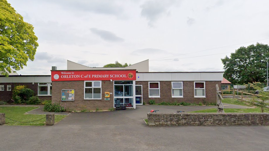 The brown brick front entrance of Orleton school. There is a red sign with the school's name on it above the front doors. The doors and windows have white panelling, and there is a small area of grass on either side of the front entrance. On the right there is a scarecrow-like statue.