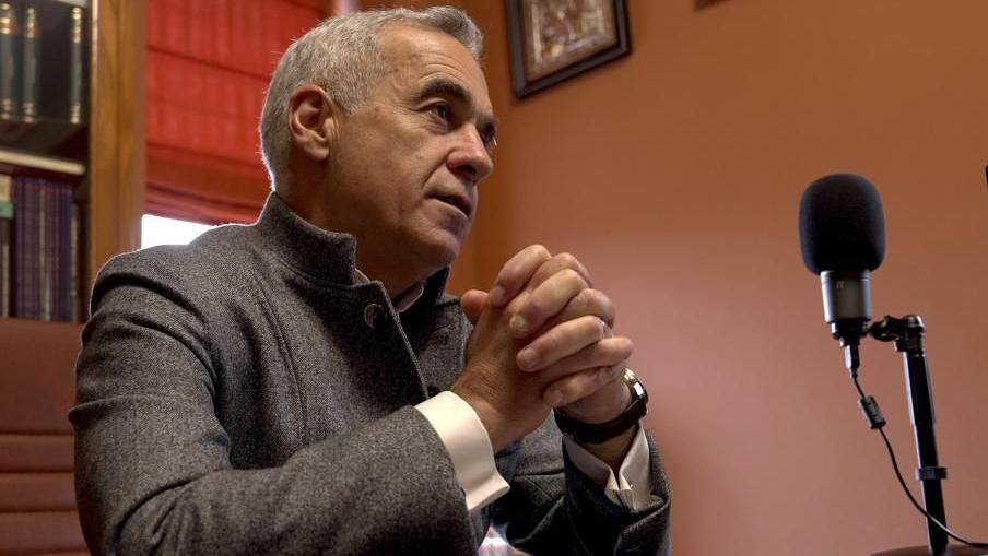 Georgescu, a man with grey hair and wearing a shirt and grey jacket, sits at a desk, his hands clasped, looking pensive. There is a single microphone in front of him.