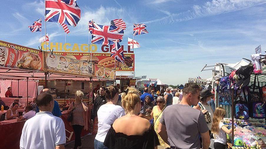 A busy market scene where people are walking between stalls. To the left is a stall with Union Jack and US flags flying above a stall selling food. To the right is a stall selling hats and sweets.