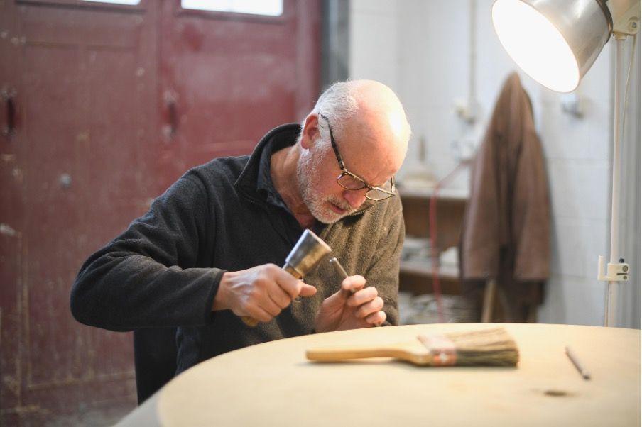 A bald man with glasses with a hammer and chisel in his hands as he works on the sculpture in a workshop under a bright lamp
