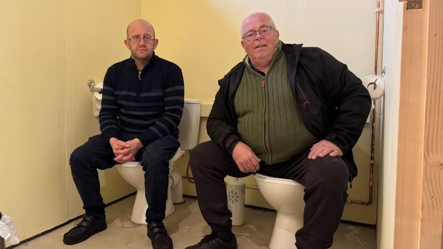 Scout leaders Peter and John Clarke sit on toilets which are situated side-by-side, but have no dividing partitions or doors. The walls are painted yellow, and there is bare wood visible on a door post.