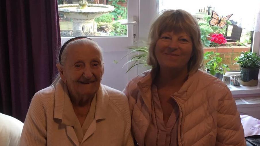 An elderly woman in a white cardigan and a younger woman in a coat and with lighter hair. They're in a room with a patio door behind, both sat on a bed.