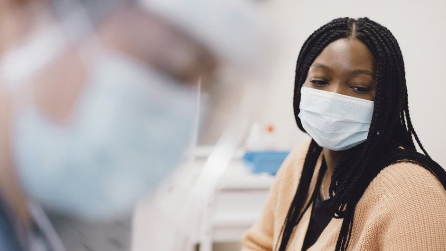 A doctor wearing a mask, blurred in the foreground with a patients, also wearing a mask, in the background