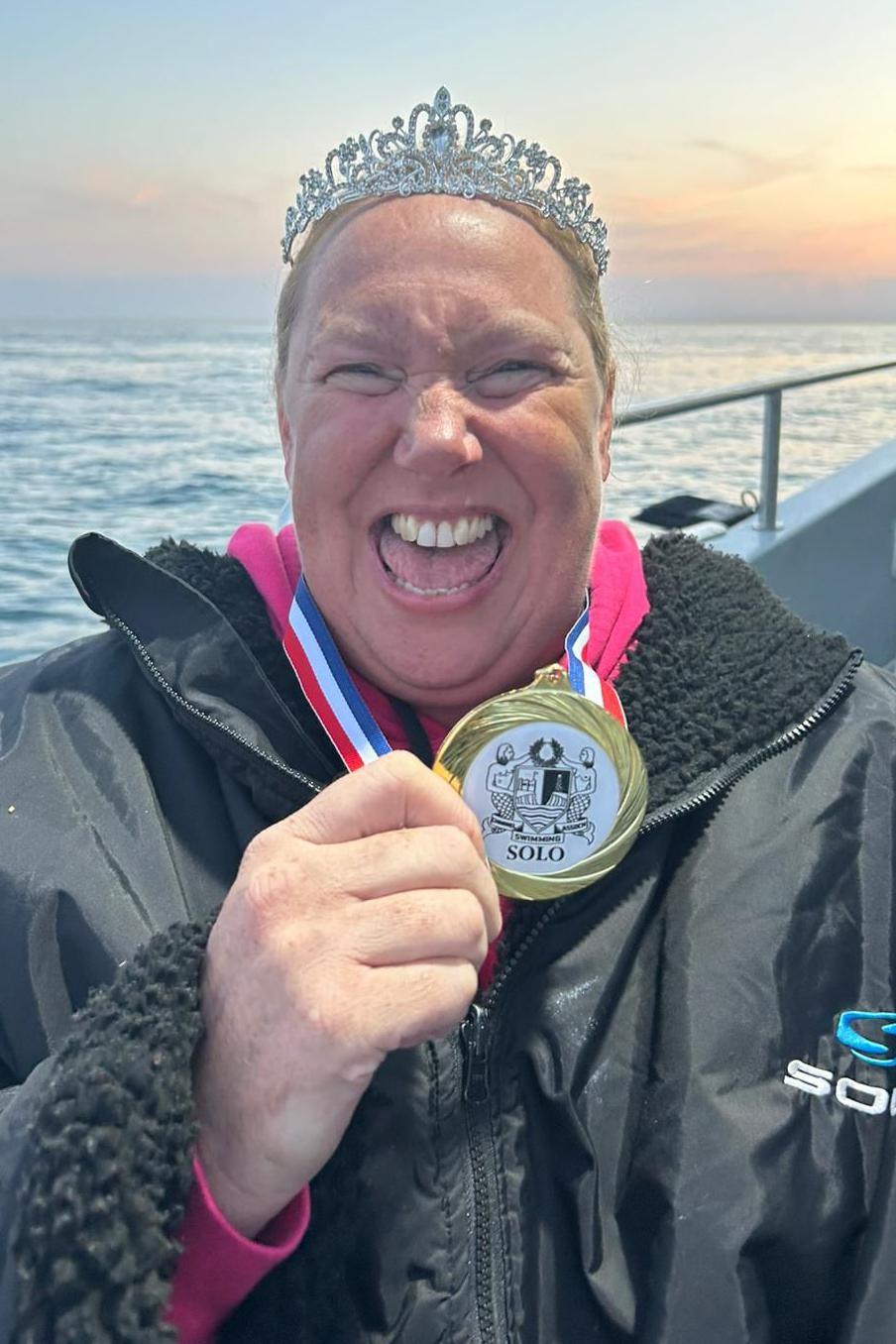 Lindsey Walke, wearing a swim robe and a tiara, she is holding her medal. She is facing the camera and smiling with her eyes squinted. Behind her is the side of the boat. The sea behind her is a light blue and the sky is a light blue to orange ombre.