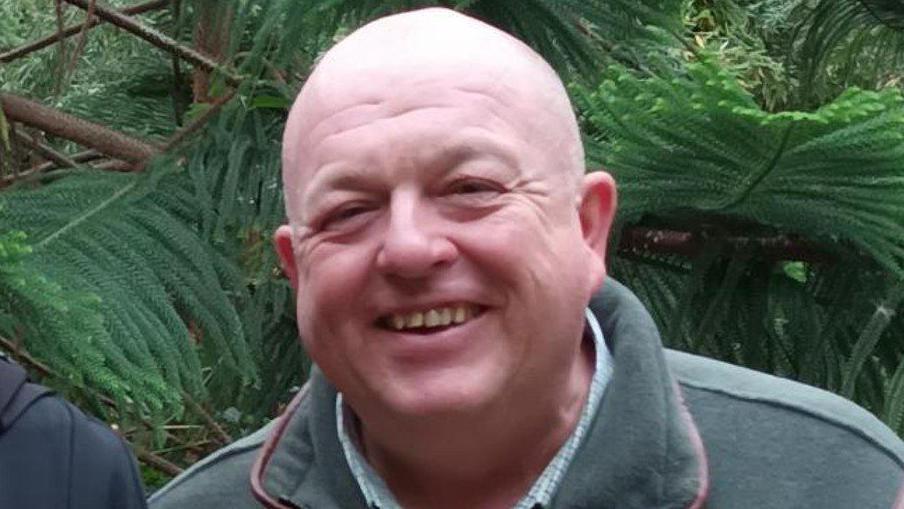 A close-up picture of Martin Bembridge. He is smiling and has a bald head and is wearing a green jumper with a shirt underneath.