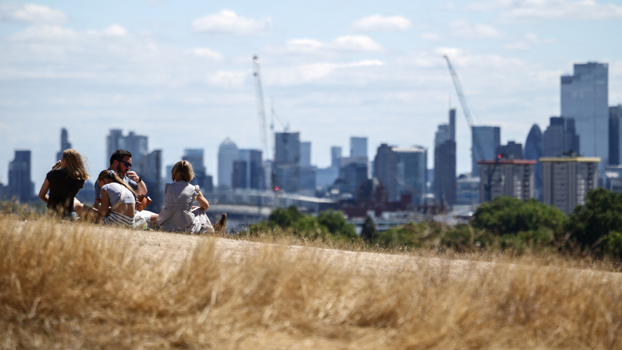 A group of people sit on the dry, sun-scorched grass of Primrose Hill with the skyline of central London in the background
