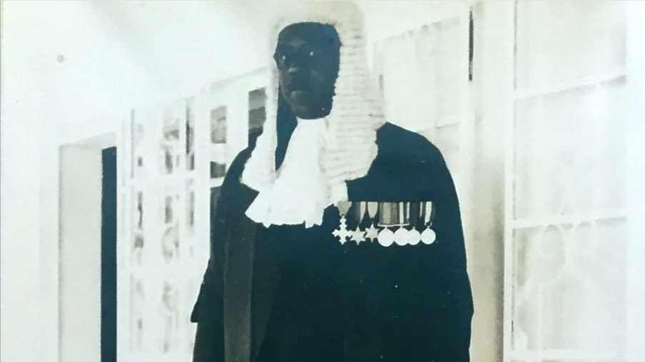 Johnny Smythe standing looking at the camera in full dress cloak and white wig as a barrister. He has all his medals on his black cloak.