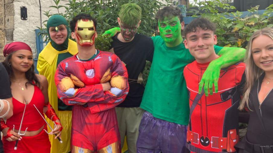 A group of young men and women wearing fancy dress costumes, including superhero outfits, masks and painted faces