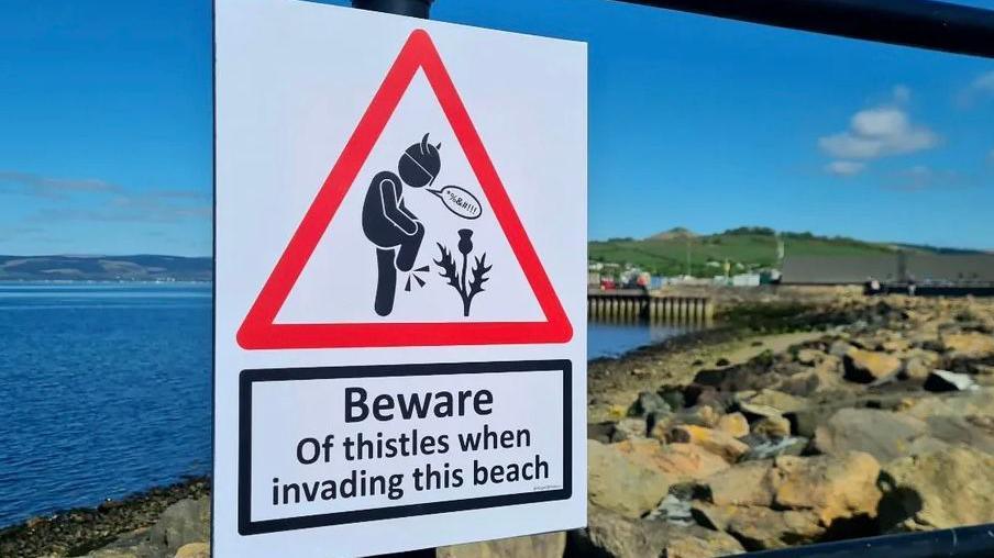 The sign has someone standing on a thistle. It says: Beware of thistles when invading this beach