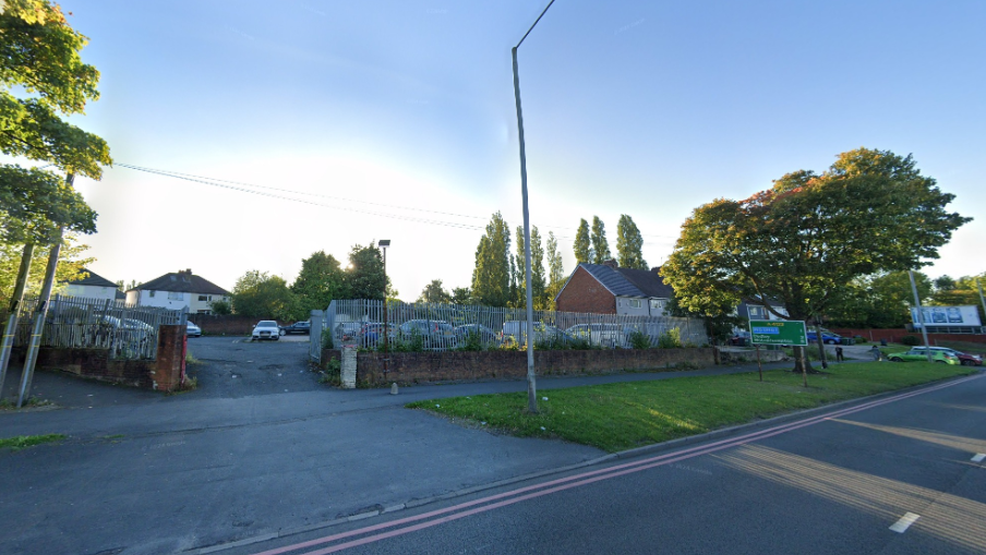 A fence surrounds the derelict land which has some cars parked on it and is set back from the road.