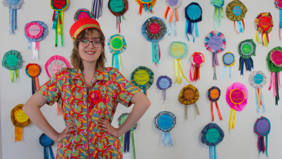 A young woman in a colourful jumpsuit wearing a red hat and glasses and with her hands on her hips, standing in front of wall covered with lots of colourful rosettes