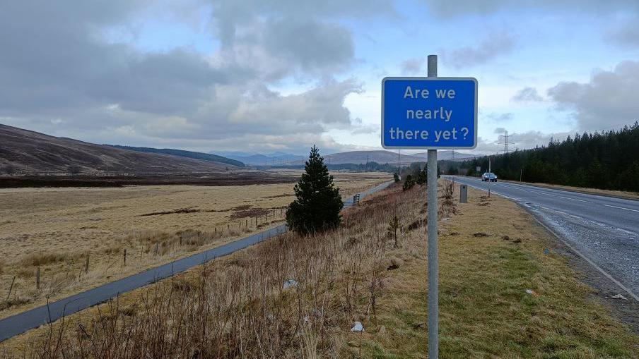 A blue road sign which reads 'are we nearly there yet' in front of a landscape view of a field and hills