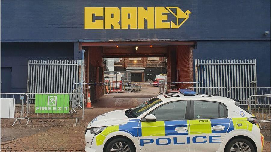 A police car parked outside a blue building with a square archway. The word crane is in yellow capital letters in the centre of the building