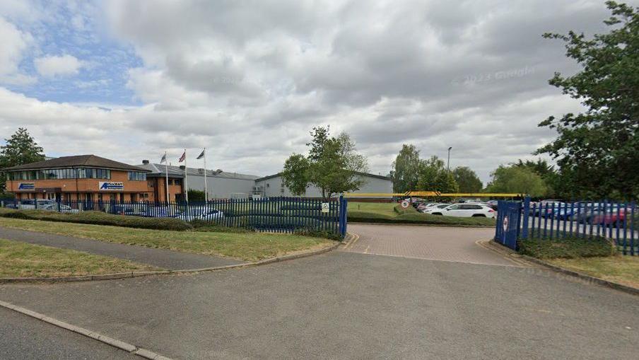 An industrial area with a section housing several businesses, enclosed by blue fencing and a yellow barrier. There is a two-storey brick building to the left and warehouses beyond. There are cars parked around the section, and some trees.