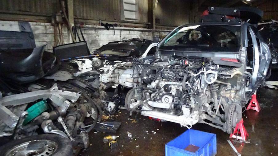 Another stripped vehicle in a garage. Just the chasis remains. The garage is piled high with car parts.