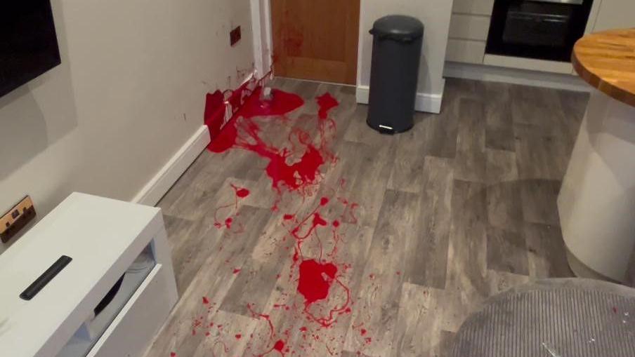 Red paint splattered across grey wood flooring and over white skirting boards and walls. 