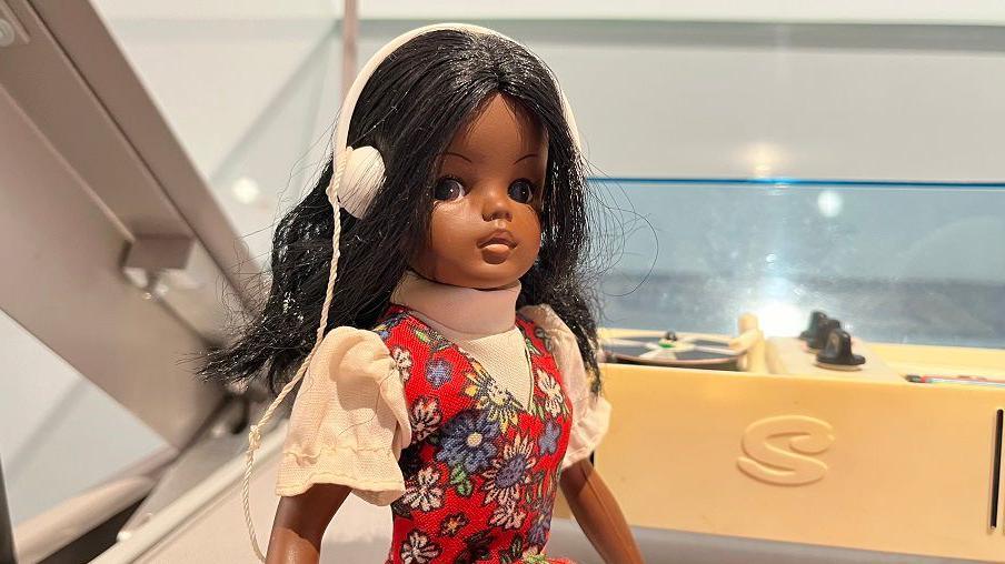 A black doll with the same shape as Sindy in a red dress and white shirt