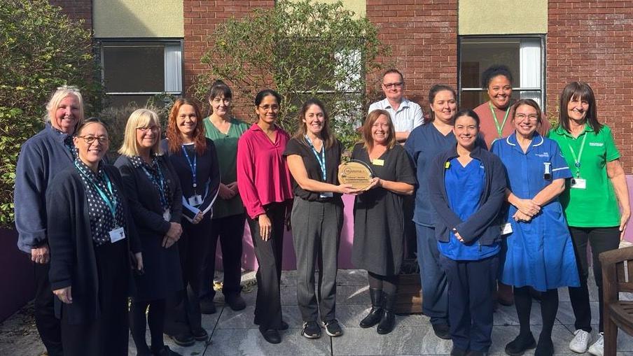 Fourteen medical staff at the Christie Cancer Centre stand outside the Tameside Hospital with two women in the centre holding a trophy on a dry day. All staff are smiling.