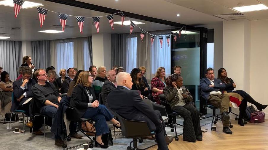 Businessmen and women are seated on chairs watching a screen as the US election results come in with bunting showing the American flag pattern above them.