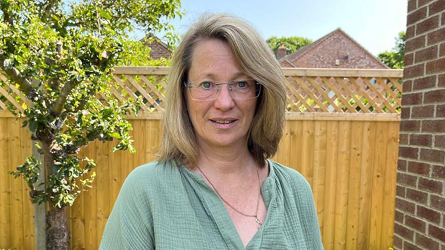 Christine Cullen, wearing a green blouse and a chain around her neck, stands in a garden. She is wearing glasses and has shoulder-length blonde hair. Behind her is a wooden fence and a tree.